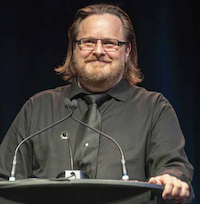 A bearded man wearing glasses and a black shirt and tie, addresses an off-camera crowd from a podium.