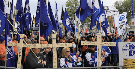 A large crowd of people carrying blue M&eacute;tallos flags and three wooden crosses protest behind a barricade outside the Quebec National Assembly.