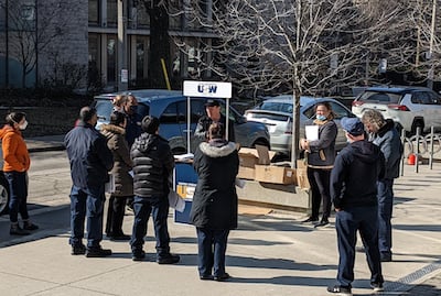 A group of spaced-out people in coats and masks listen to a man speaking from a pop-up booth with a USW logo on it. There are trees and parked cars in the background. 