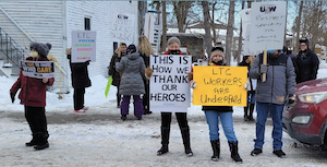 A group of 15 people, mostly women, wearing toques and heavy winter coats, hold placards and picket signs on a town street. There is snow on the ground.
