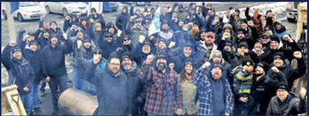 A group of 50 or more people, in dark winter coats, with hats, raise their fists and smile for the camera - solidarity on the picket line in Contrecoeur, Quebec