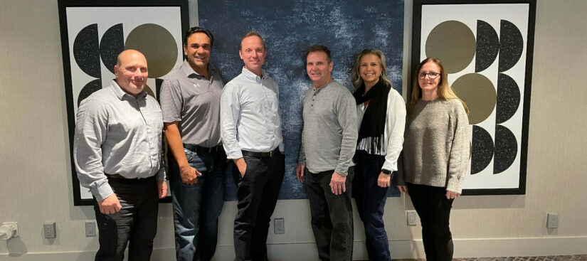 Image: A group of six people together. They are inside and posing. They are wearing dark pants and light and grey coloured tops.