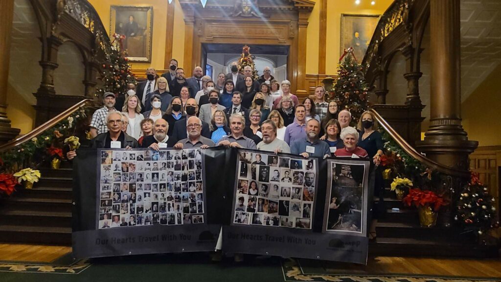 Rows of people standing on the large staircase in the legislature. There are 7 people at the very front holding a banner with photos of various miners who were affected by McIntyre Powder. Text at the bottom of the banner says "Our hearts travel with you"