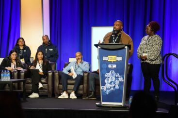 Image: A Black man stands at a podium speaking into a microphone with a Black woman standing standing on his left. To his right, four people sit in armchairs, and two people sit on stools behind the row of armchairs.