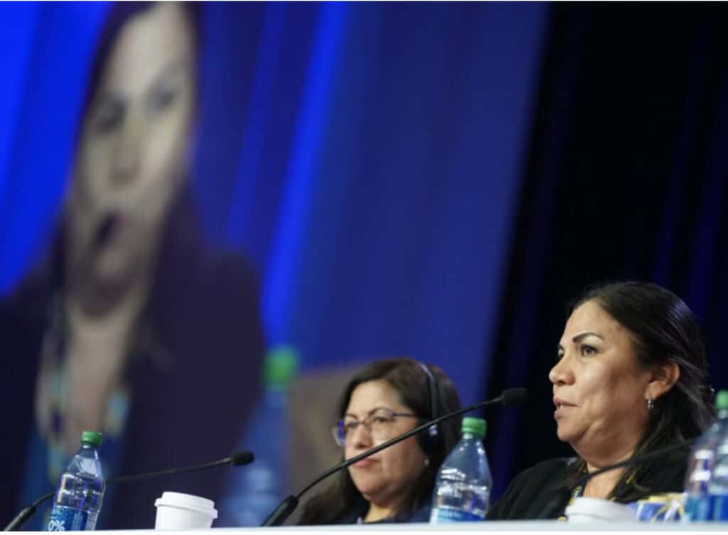 Image: Two women are in the lower-right of the frame, seated with microphones on the table before them. A blurred-out projection of the speakers on a large screen is in the background behind them.