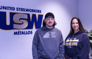 Image: A man and a woman wearing sweatshirts stand in a reception area facing the camera. Behind them is a giant USW logo on the wall of the space.