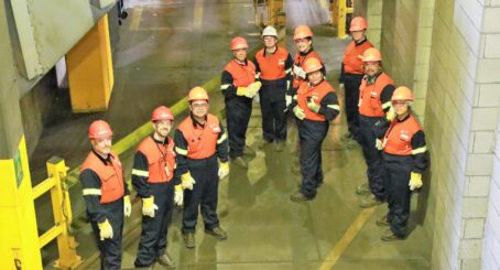 Image: Seen from above, a group of 10 people, all wearing orange hardhats (except one woman in a white hardhat), black coveralls and orange safety vests and yellow gloves are looking towards the camera while touring an industrial workplace