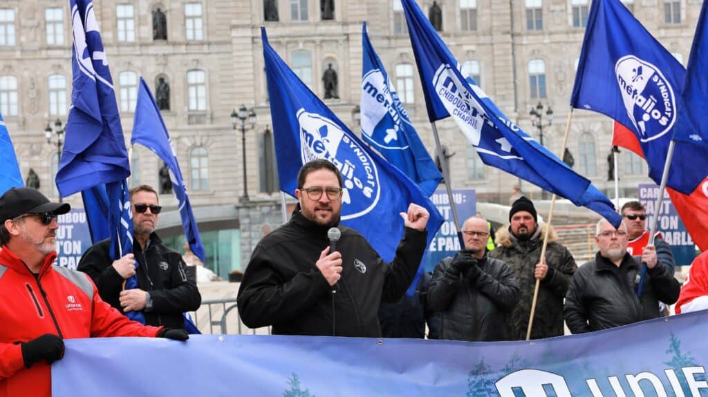 Image: A man stands behind a banner with numerous blue-and-white flags behind him. He is joined by six other people. They are outdoors. The man is speaking into a handheld microphone.