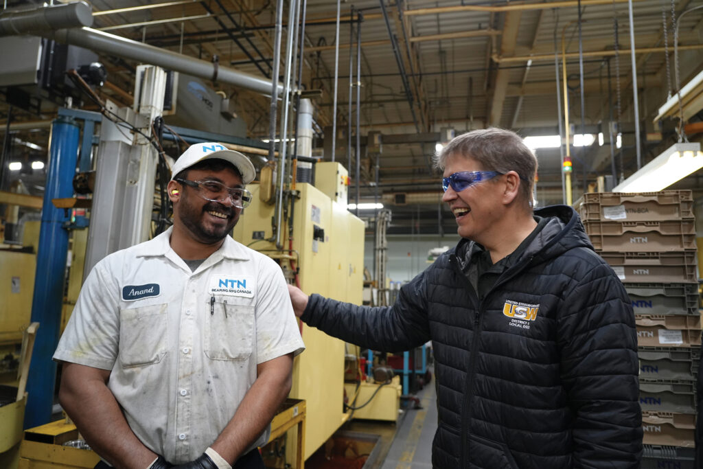 Image: a Black man and a white man greet each other, smiling, in an industrial setting. The Black man is wearing a work uniform with "NTN" above the shirt pocket. He is wearing a white ballcap. The white man is wearing a dark puffy jacket with USW on it.