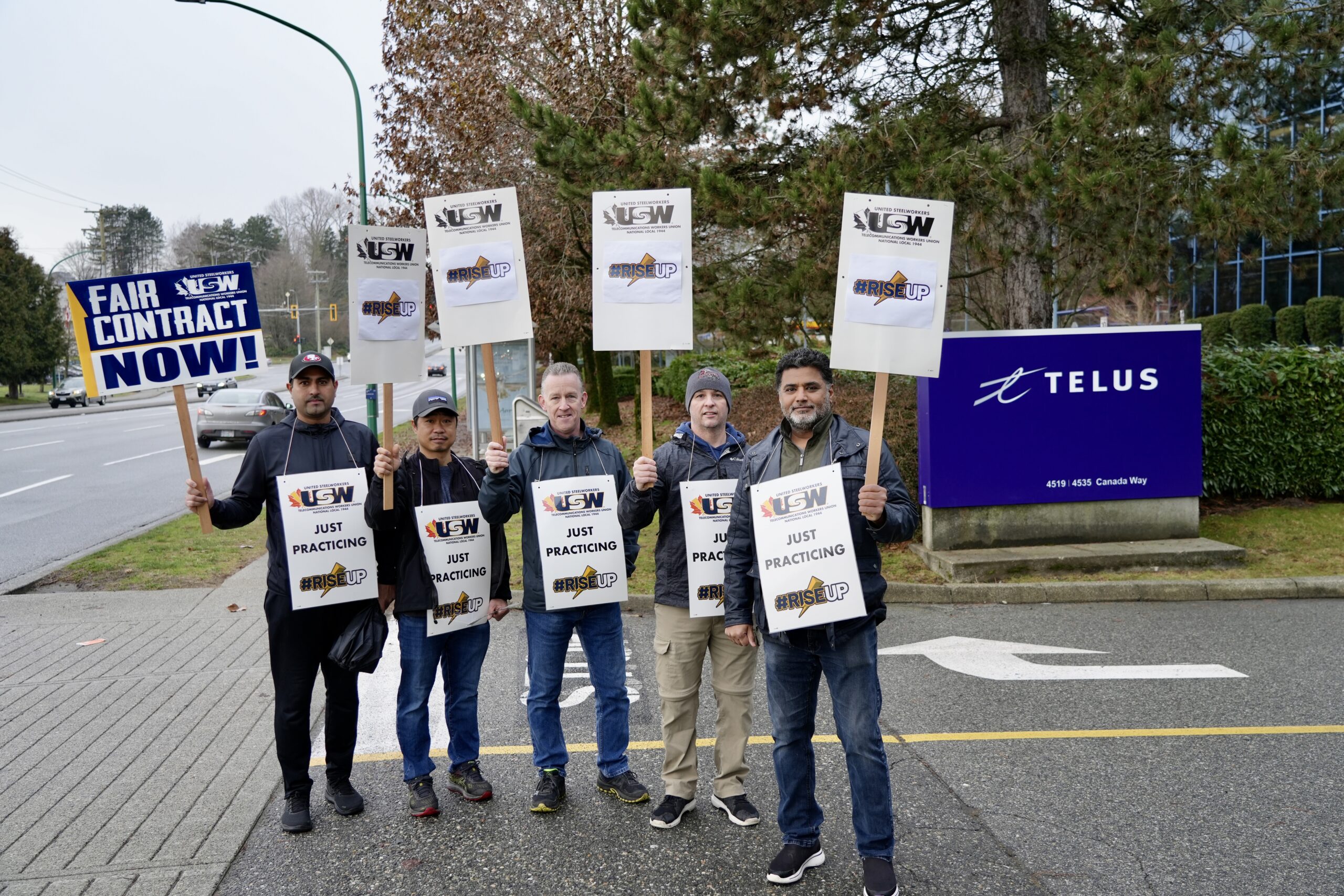 Image: a group of five mean wearing picket signs and holding up signs stand outdoors in the road outside a Telus building. Signs read: Fair Contract Now and Just Praticing.