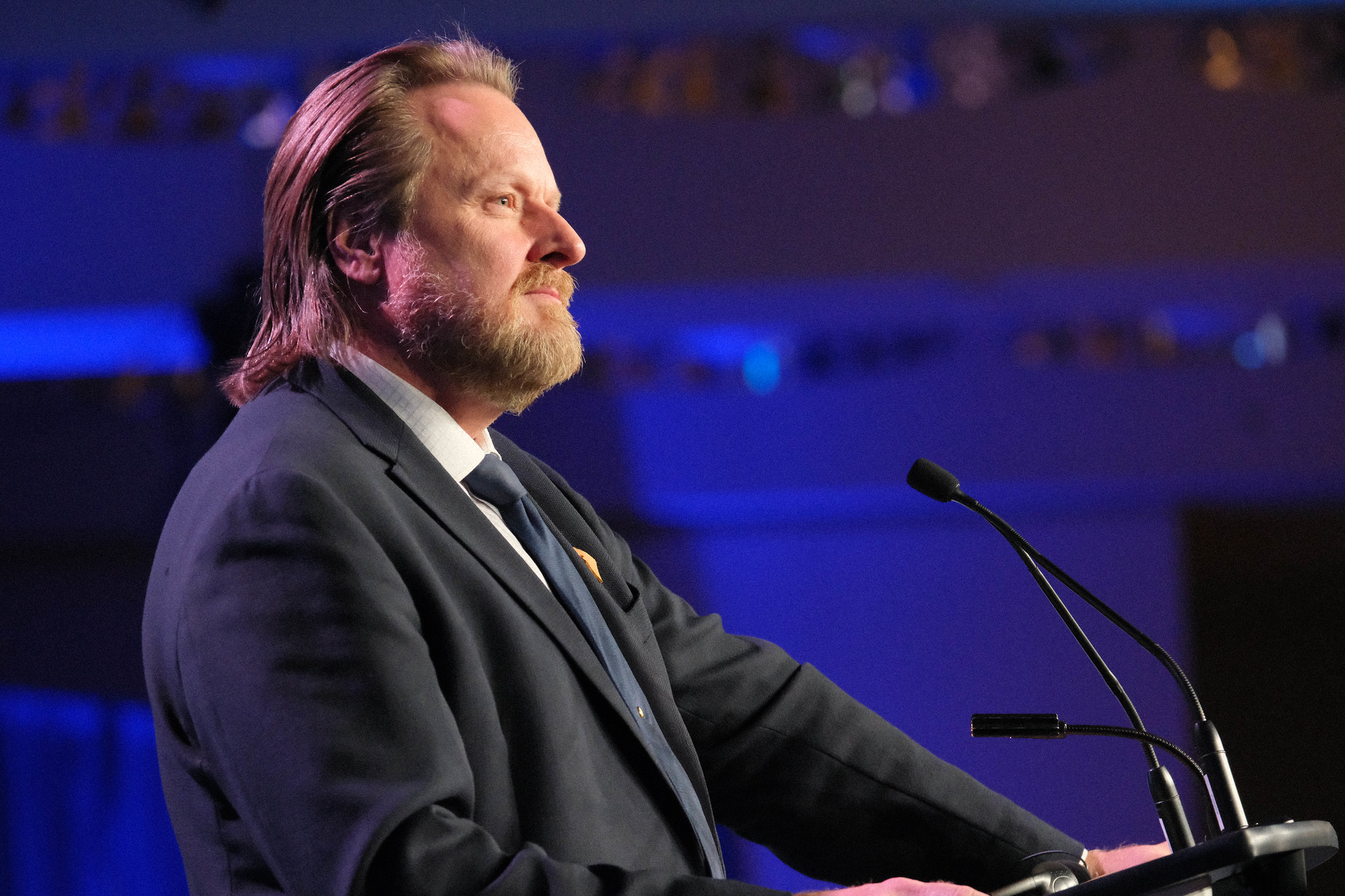 Image: profile image of a bearded man in a suit and tie standing at a podium with microphones looking out into an audience outside the frame. There is a wash of royal blue lighting in the background.