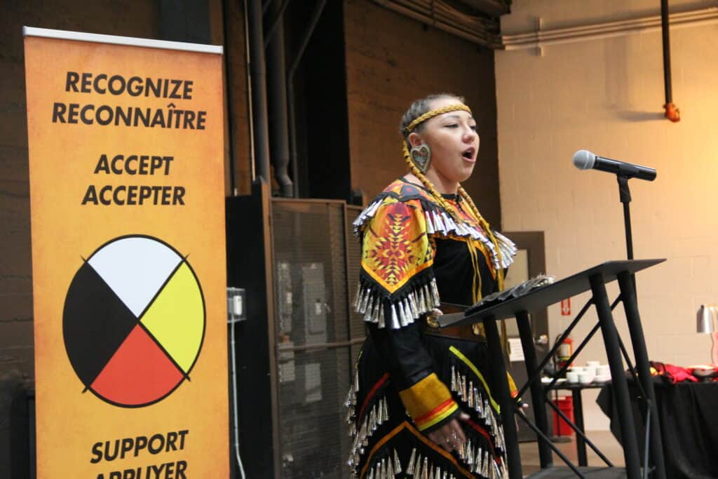 Image: a young woman in traditional Indigenous clothing sings at a microphone. Behind her is a stand-up banner with text: Recognize Reconnaître, Accept Accepter, Support Appuyer as well as an Indigenous medicine wheel symbol