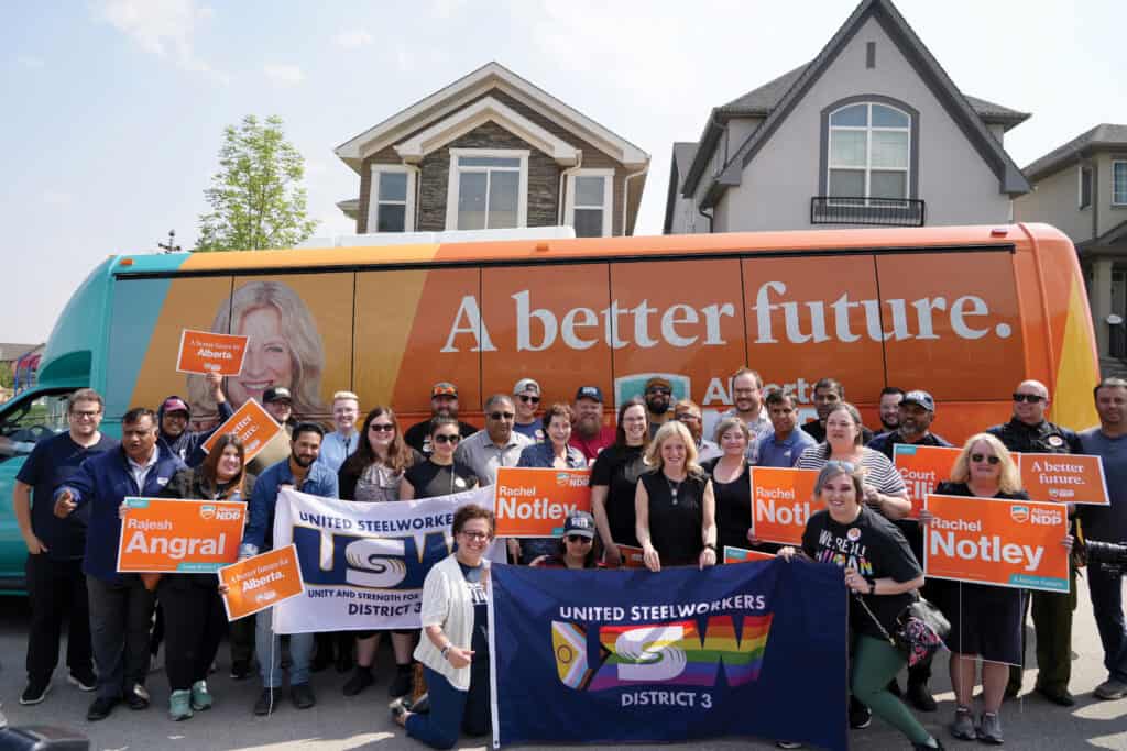 Image: A group of smiling people holding USW flags and orange election signs, stand with a woman (Rachel Notley) in front of an orange bus with text: A better future