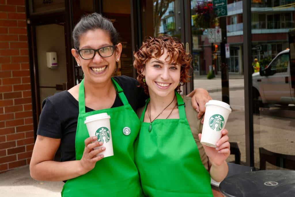 Two partners from Starbucks wearing green aprons smiling and holding coffee cups