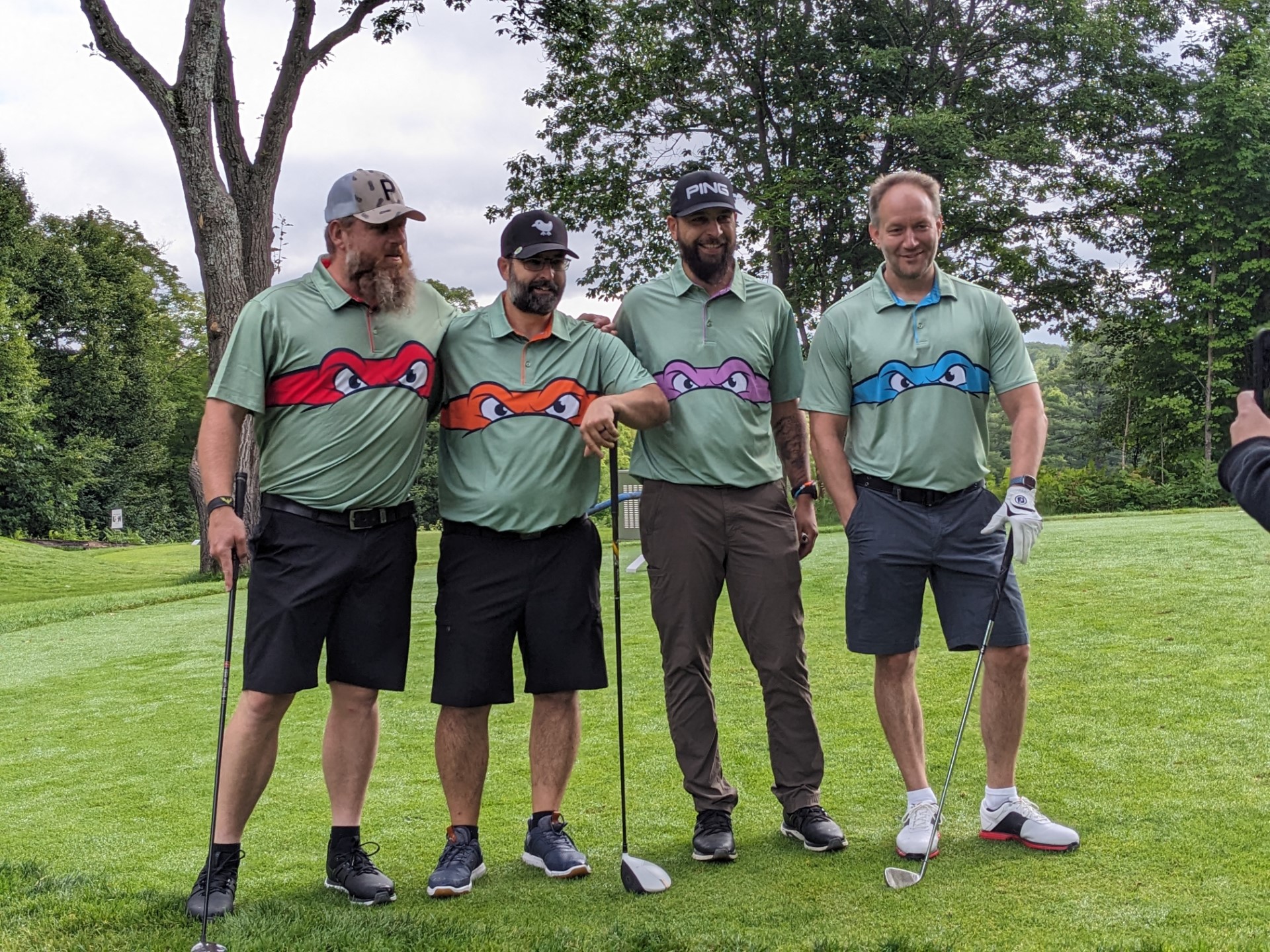 Four people standing side by side, outdoors. The individuals first and second from the left and the individual on the far right are holding golf clubs.