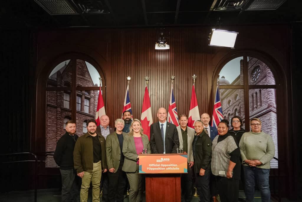 An image of a group of people standing side-by-side indoors. There is a podium in front of the person standing in the middle of the group. Behind them are 6 flags draping on flagpoles.