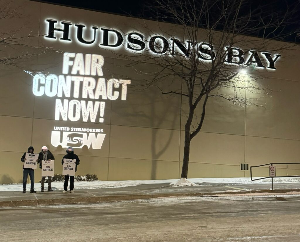 "Fair Contract Now" is projected on a Hudson's Bay Store in Kamloops with 3 striking workers standing in front.