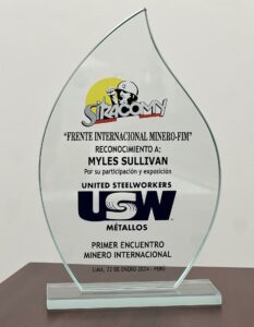 A image of a glass plaque shaped like a flame. There is a logo at the very top of a miner with a fist up and the acronym of the union SIRACOMY. Underneath is text in Spanish that says "Frente Internacional Minero-Fim reconocimiento a: Myles Sullivan por su participación y exposición primer encuentro minero internacional. Lima, 22 de enero 2024 - Peru' There is also a United Steelworkers/USW Métallos logo on the plaque.