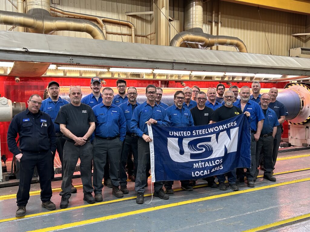 An image of people inside a manufacturing plant. The group is standing close, side-by-side and there is a blue flag in the middle that's being held up. The flag says 'United Steelworkers USW Métallos Local 6565'