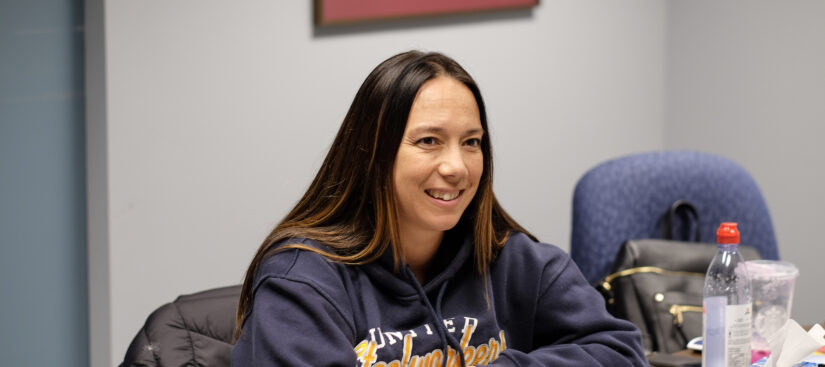 Image: A person with long hair wearing a USW sweatshirt is seated at a table with documents in front of them. They are smiling and looking across the table at someone else off-camera.