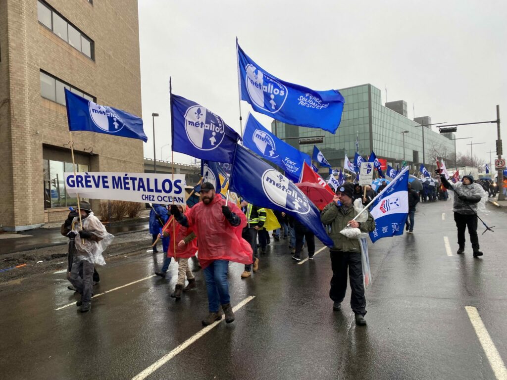 A parade of striking Steelworkers marching down a street waving, waving Métallos flags.