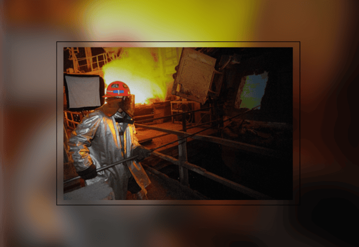 A steel worker in full safety gear working in front of a blast furnace.