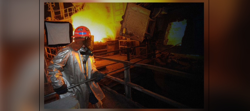 A steel worker in full safety gear working in front of a blast furnace.