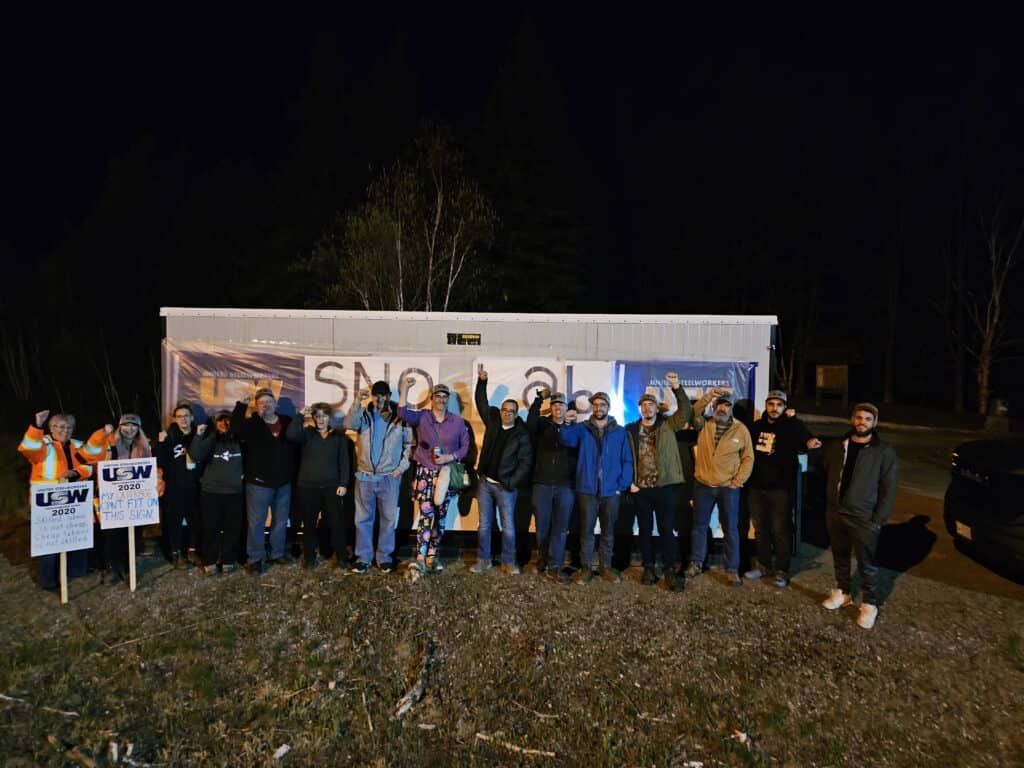 A group of people standing side-by-side outdoors, in front of a trailer. The photo was taken in the evening, marking the beginning of a strike, so the surroundings are dark.