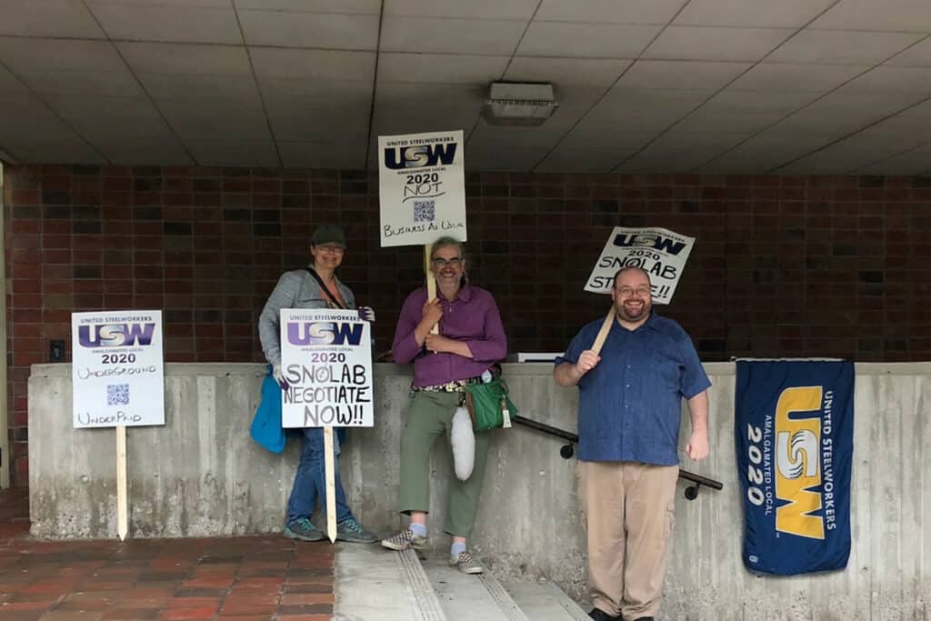 An image of three people standing outside of a building, holding picket signs. There is a USW flag draping off the knee wall.