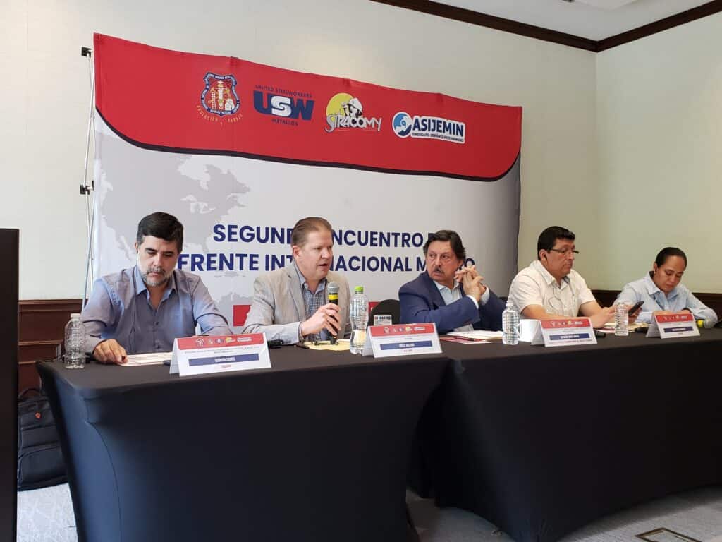 A photo of 5 people sitting behind a table. There is a banner red and white banner behind them with logos of 4 unions. 