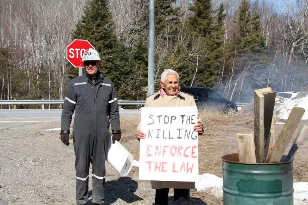 Image: Two people standing outdoors. The person on the left is wearing a grey work jumpsuit and safety helmet, and the person on the right is wearing a beige winter jacket and holding a placard with the message saying “Stop the killing, enforce the law.” There is a metal barrel next to them with smoke and pieces of wood coming out of it. A black car and numerous trees are in the background.