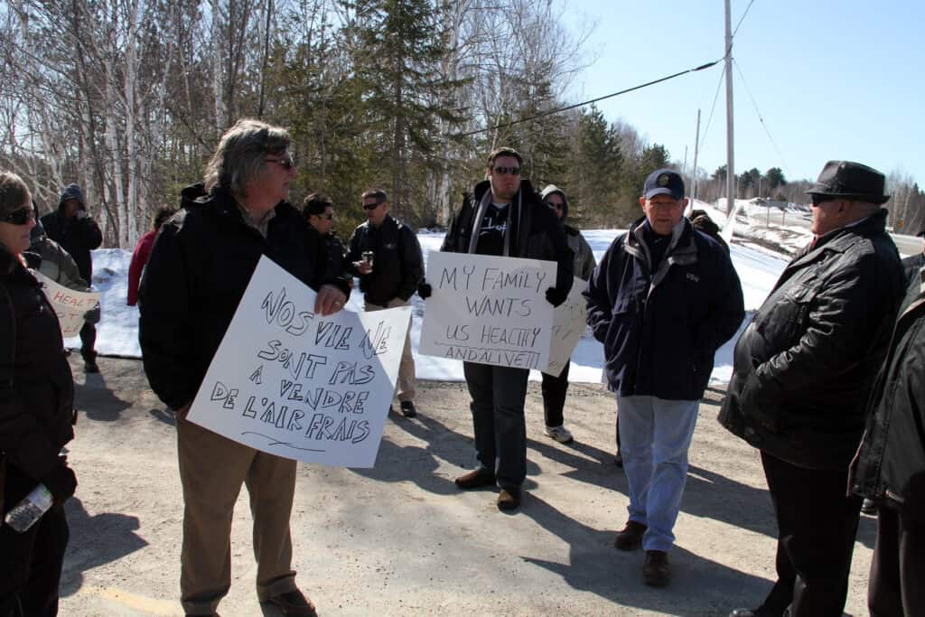 Image: Several people wearing winter clothes attending a picket line and holding placards with various messages. Two placards are visible at the front, one in French saying “Nos vies ne sont pas à vendre, de l’air frais,” and the other in English saying “My family wants us healthy and alive.” Trees and a snow-covered area are in the background.