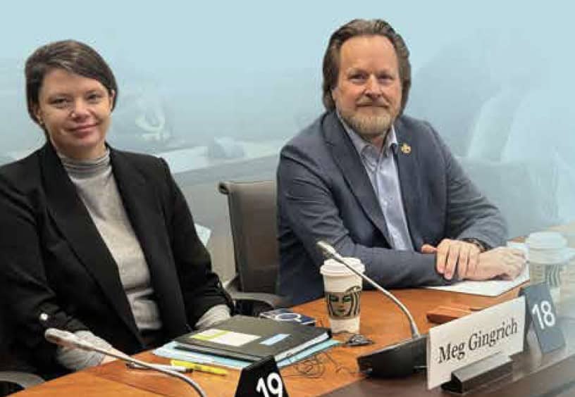 Image: Two people sitting in a boardroom, looking at the camera for a photo. The person on the right is wearing a grey suit with a blue shirt, and the person on the left is wearing a black blazer and grey sweater.