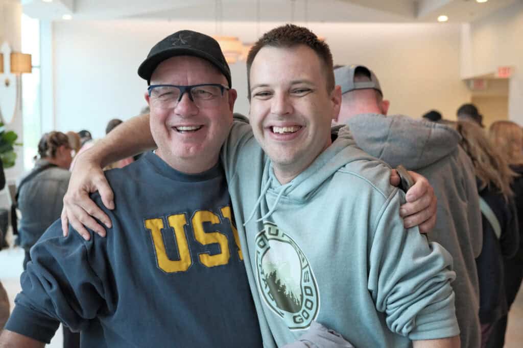 Image : two smiling people facing the camera wearing sweatshirts, one with an arm around the other in a friendly fashion.