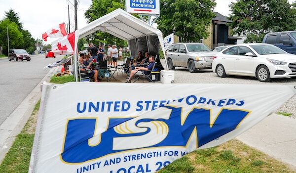 Image: A USW/Métallos Local 2918 flag on the foreground. In the background is a tent with people sitting on chairs inside it.