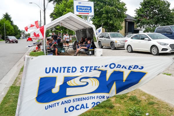 Image: A USW/Métallos Local 2918 flag on the foreground. In the background is a tent with people sitting on chairs inside it.