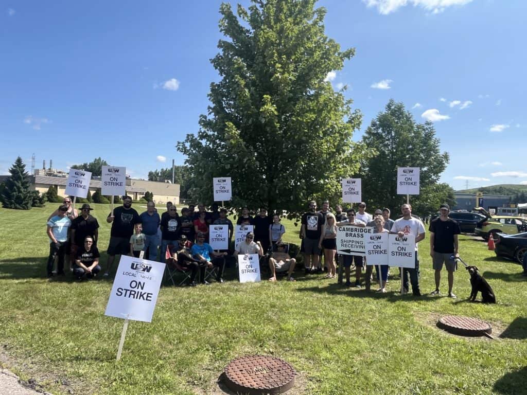 An image of a group of about 30 people standing side-by-side outdoors. There are various picket signs either staked on the lawn or being held up that say 'On Strike.'