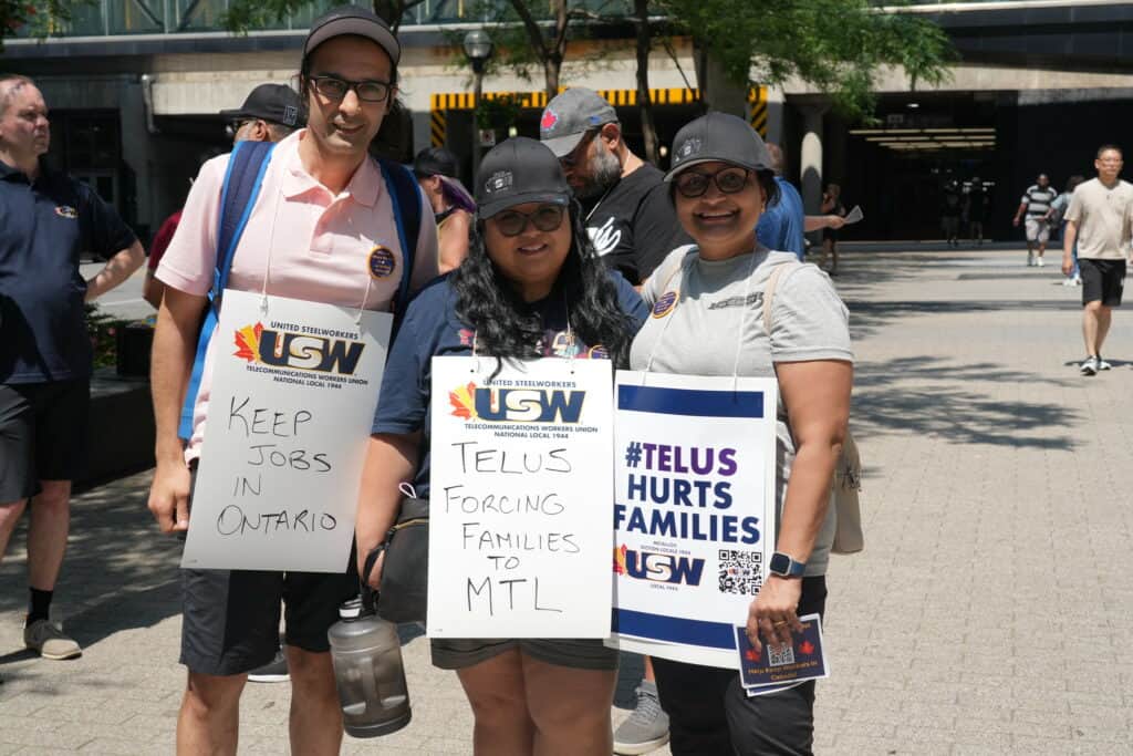 Image: three people are facing the camera, outdoors on a sunny day. They are wearing placards reading "Keep jobs in Ontario," and "#Telus Hurts Families."