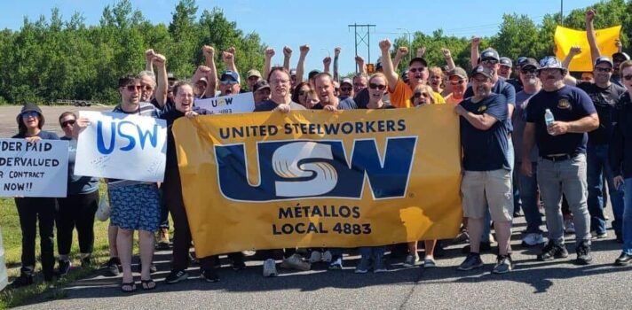 A group of people standing in the street during a rally pumping fists and holding USW flags and signs.