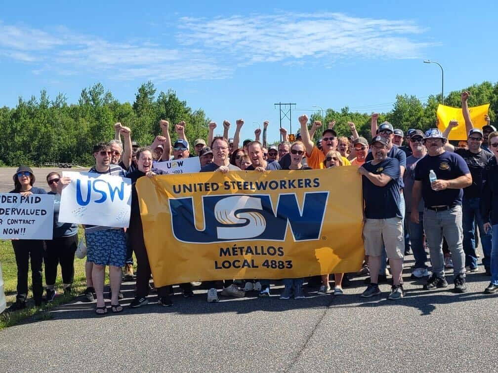 A group of people standing in the street during a rally pumping fists and holding USW flags and signs.