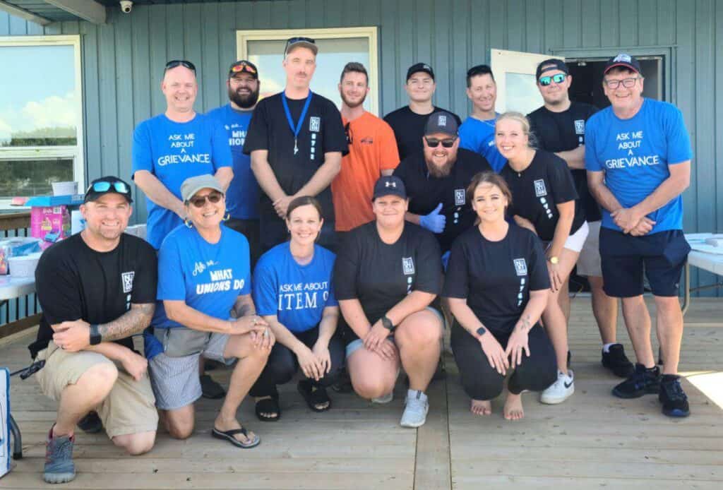 Image: a tiered photo of a group of more than 10 people facing the camera, some of whom are wearing blue T-shirts with "ask me about" on them