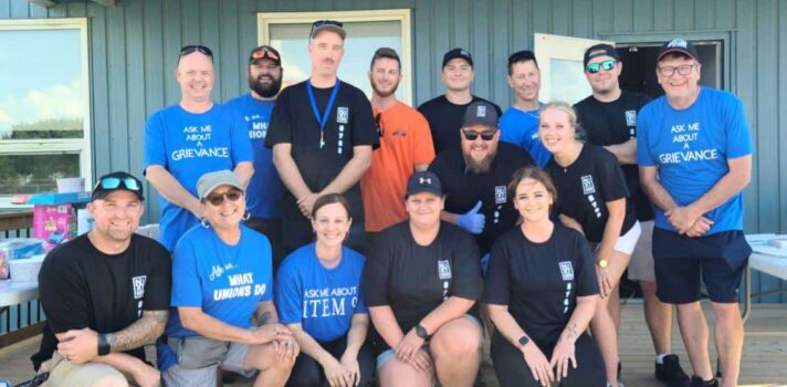 Image: a tiered photo of a group of more than 10 people facing the camera, some of whom are wearing blue T-shirts with 