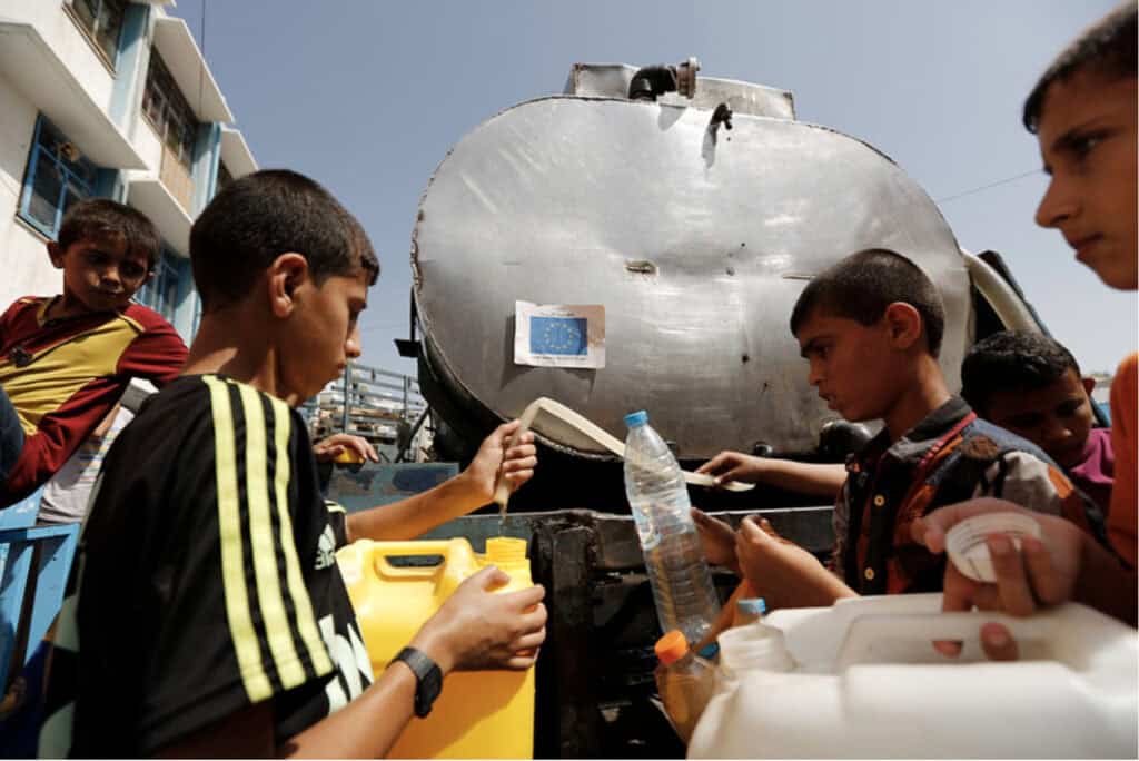 A group of kids holding empty water gallons and lining up in front of a water container waiting for their turns.