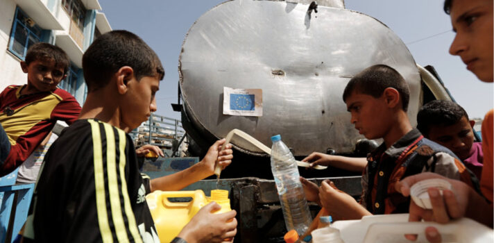 A group of kids holding empty water gallons and lining up in front of a water container waiting for their turns.