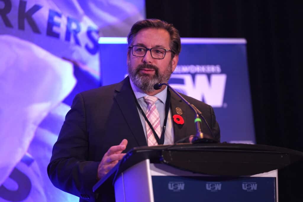 Image: A bearded man wearing glasses is at a microphone at a podium. A portion of a large screen is visible behind him with part of a United Steelworkers flag visible.