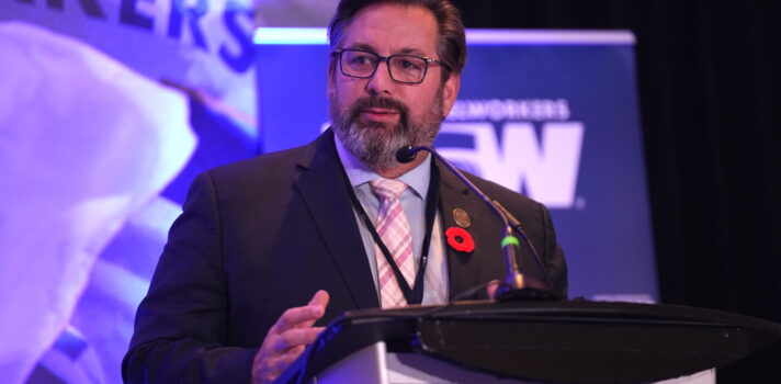 Image: A bearded man wearing glasses is at a microphone at a podium. A portion of a large screen is visible behind him with part of a United Steelworkers flag visible.