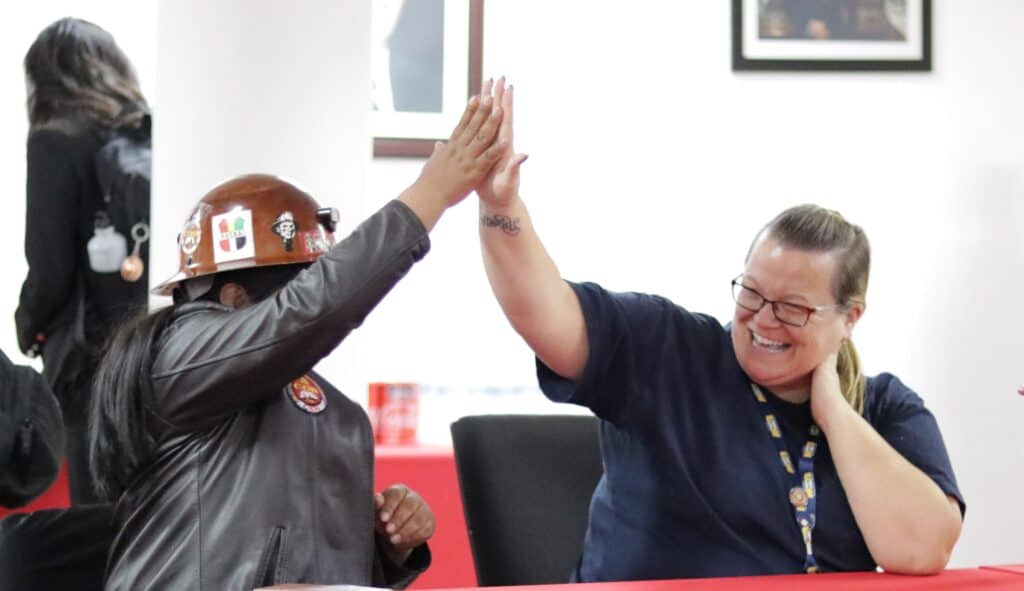 Two individuals giving each other a high five while sitting behind a table in a room. One of them is wearing safety helmet.