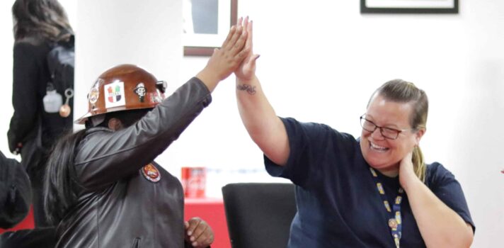 Two individuals giving each other a high five while sitting behind a table in a room. One of them is wearing safety helmet.