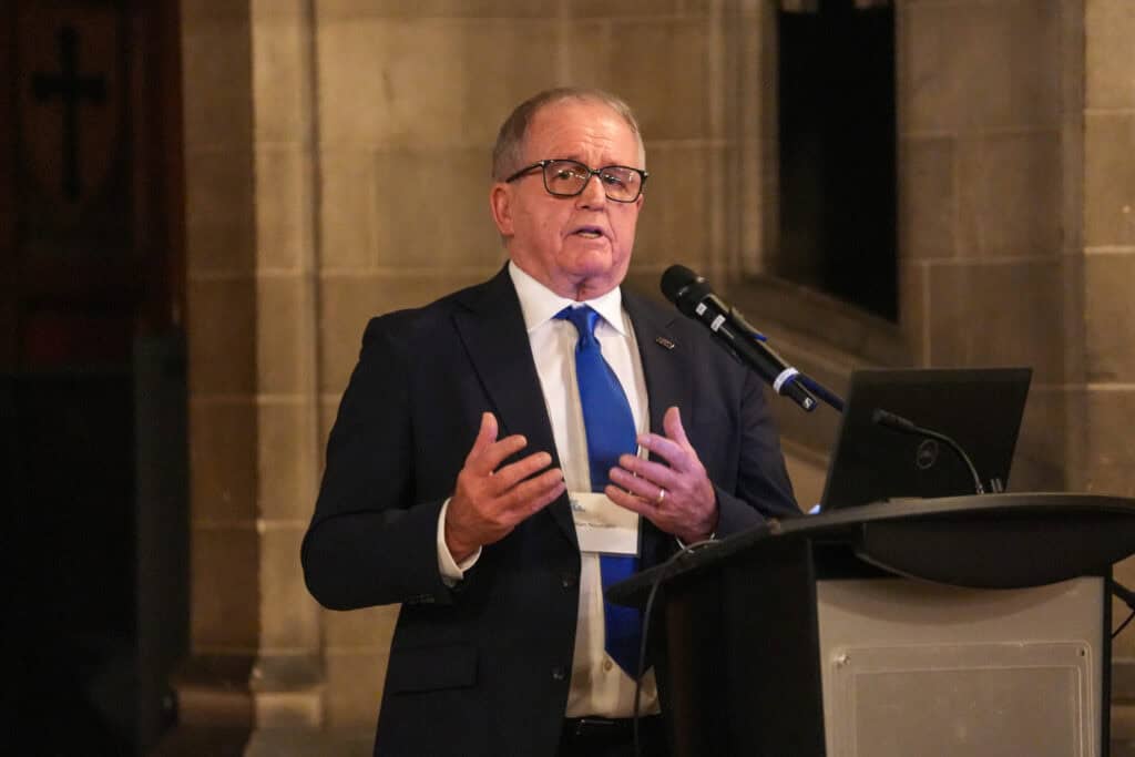 A man in a suit stands in front of a podium with a laptop on it and a microphone mounted to it. He's speaking with his hands up at chest-level as he gestures.