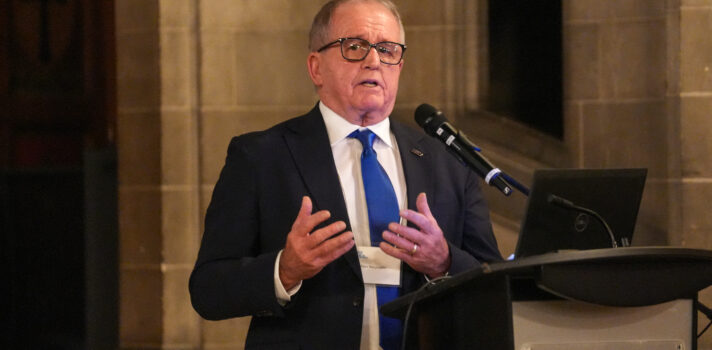 A man in a suit stands in front of a podium with a laptop on it and a microphone mounted to it. He's speaking with his hands up at chest-level as he gestures.